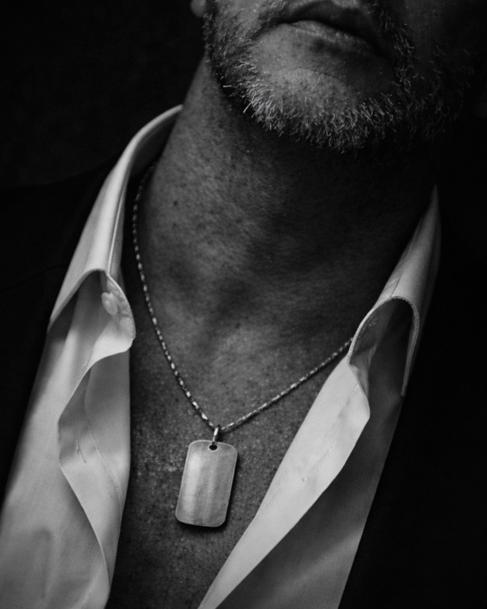 Black and white close-up of man in open shirt wearing sterling silver dog tag necklace, minimalist and symbolic MARKED jewellery aesthetic