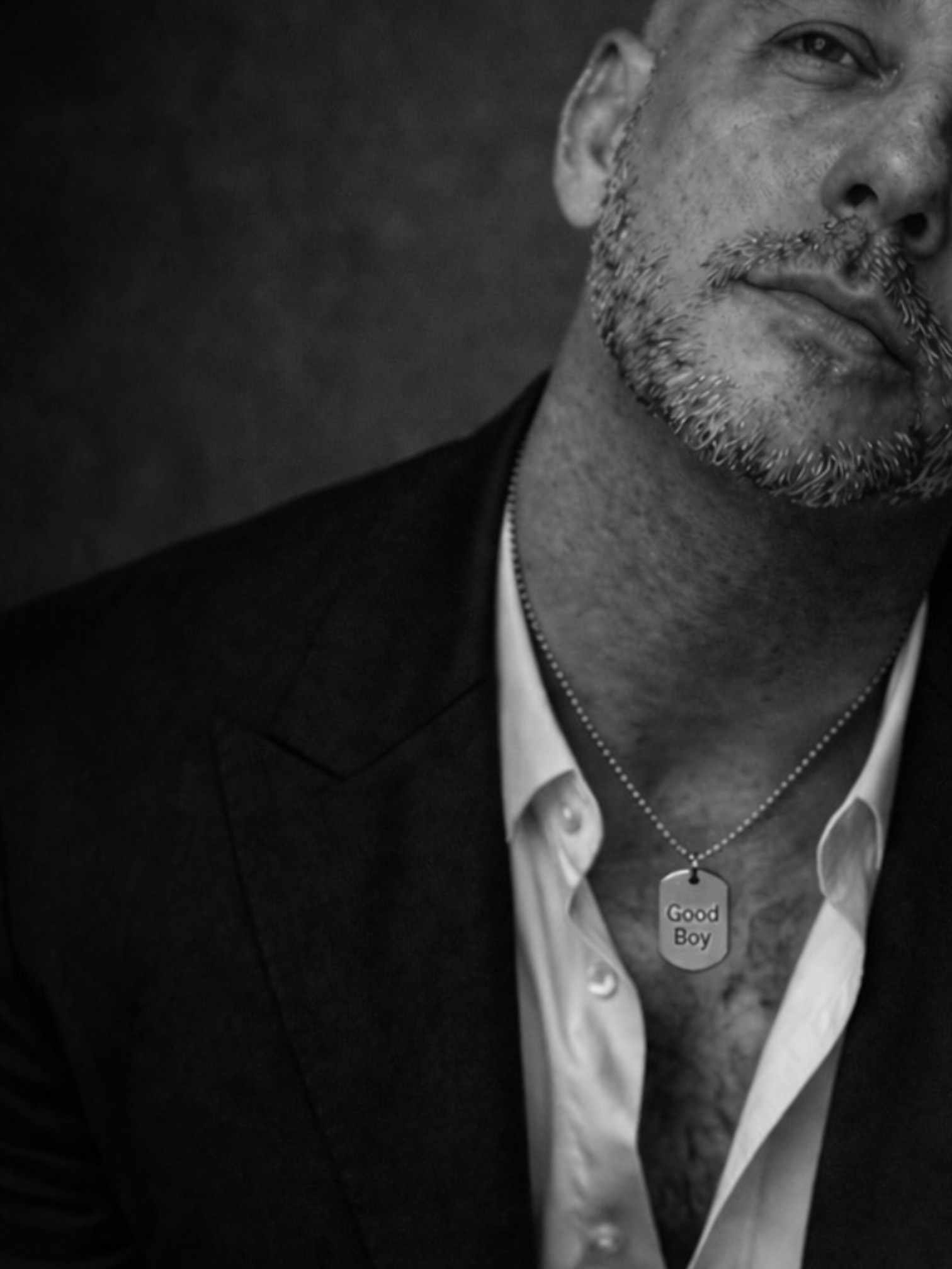Black and white portrait of man in open white shirt wearing a subtle dog tag necklace engraved Good Boy, minimalist editorial style