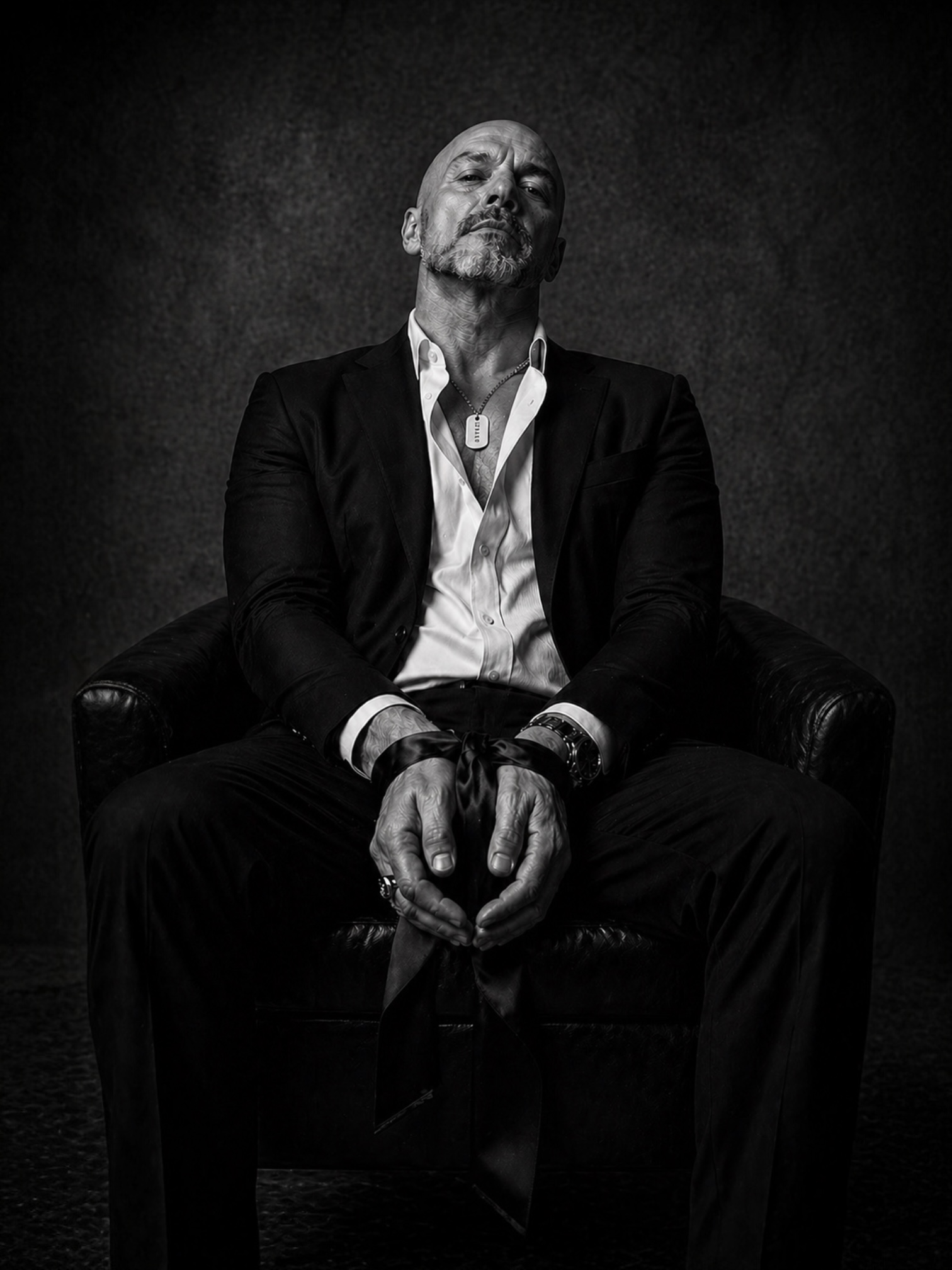 Black and white portrait of a man in tailored black jacket wearing a sterling silver MARKED dog tag, wrists softly bound with satin ribbon as a symbol of restraint, trust and control.