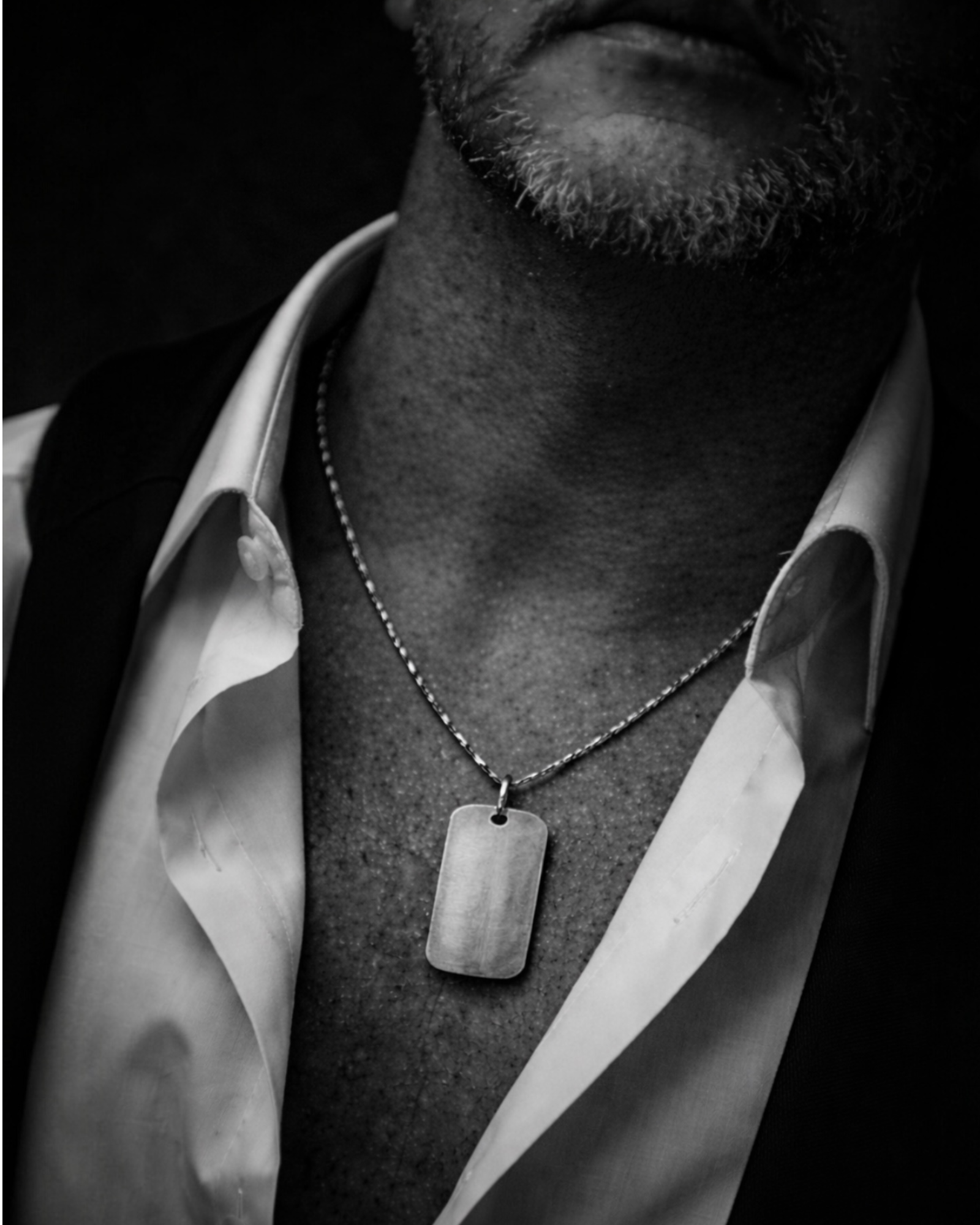 Black and white close-up of sterling silver dog tag necklace worn with open shirt
